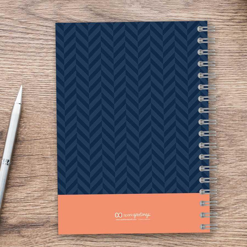 SparkGreetings® Small Anchor Writing Journal, navy blue chevron pattern cover with orange band and white logo, on wooden desk with white pen.