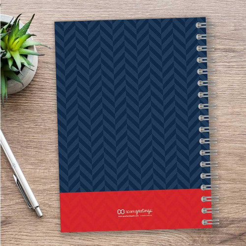 SparkGreetings® Nautical Anchor Writing Journal, navy blue cover with a red bottom section featuring a white logo and text, spiral-bound notebook on a wooden desk with a pen and a small green plant nearby.