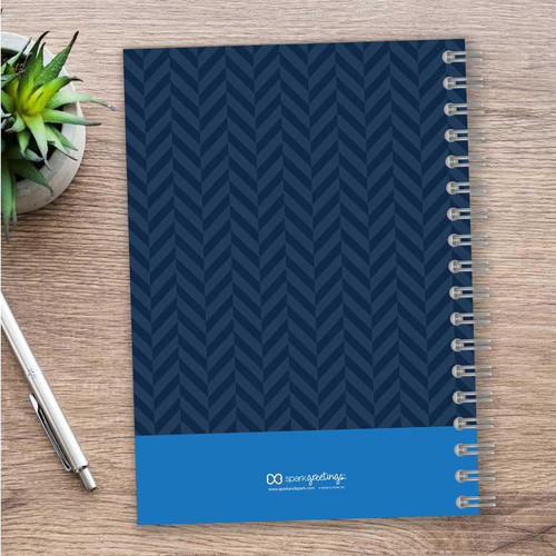 SparkGreetings® Nautical Anchor Writing Journal, navy blue cover with a chevron pattern and a solid blue band at the bottom featuring the SparkGreetings logo, on a wooden desk with a silver pen and a small potted succulent plant.