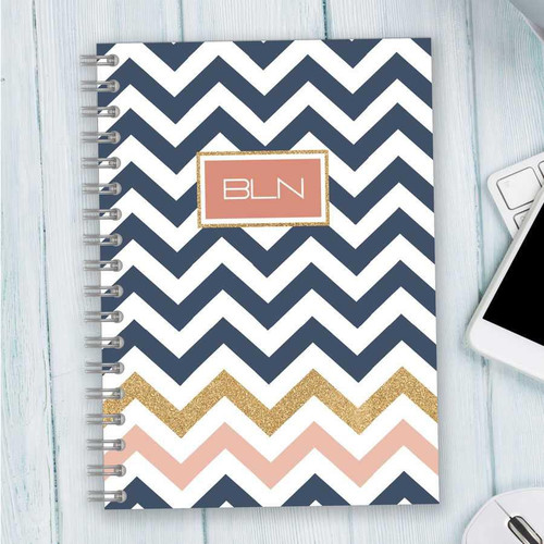 SparkGreetings® Blue Chevron Writing Journal, stylish notebook with a navy blue and white chevron pattern, gold glitter accents, pink and gold monogram highlighting space, on a light wooden desk with a tablet and computer mouse.
