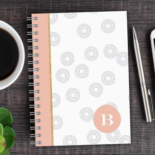 SparkGreetings® Spinning Circles Writing Journal, stylish spiral-bound journal with a white cover featuring gray circular patterns, pink accent stripe, and a monogrammed letter B in a peach circle, placed on a wooden desk with a pen, coffee cup, and phone nearby.