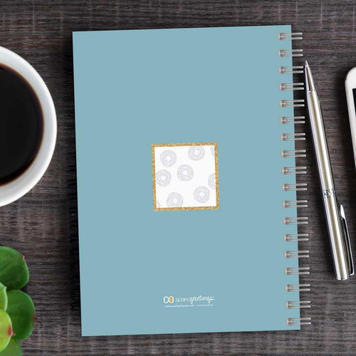 SparkGreetings® Spinning Circles Writing Journal, light blue spiral notebook with a decorative white square center featuring gray circles, on a wooden desk with a silver pen, coffee cup, and green succulent plant.