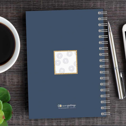 SparkGreetings® Spinning Circles Writing Journal, blue spiral-bound notebook with a white and gold patterned square on the cover, placed on a wooden desk with a cup of coffee, a silver pen, and a smartphone nearby.