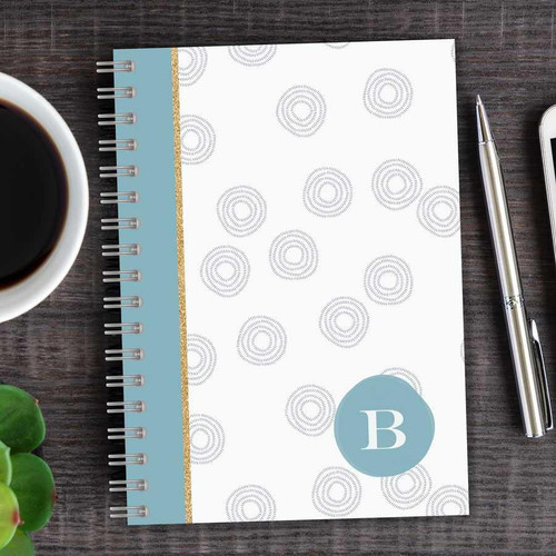 SparkGreetings® Spinning Circles Writing Journal, white journal with a blue circle and white letter B, spiral binding, decorative spirals pattern, on a wooden desk with a cup of coffee, a pen, and a smartphone.