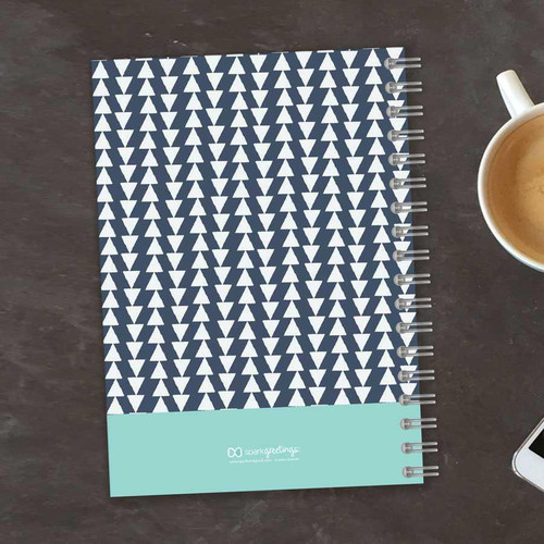 SparkGreetings® Triangles & Glitter Writing Journal, a navy blue and white geometric patterned notebook with a spiral binding, placed on a dark wooden surface next to a cup of coffee and a smartphone.