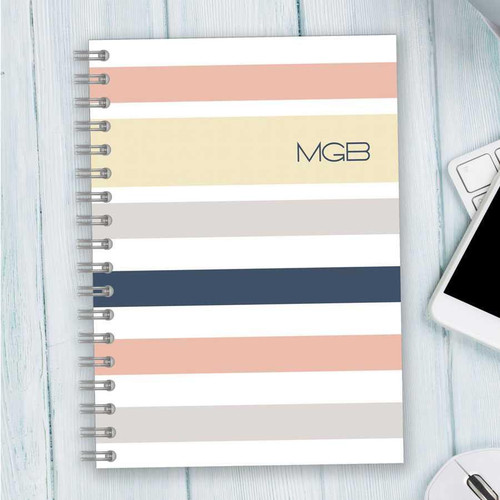 SparkGreetings® Array of Stripes Writing Journal, a spiral-bound notebook with horizontal pastel-colored stripes and the initials 'MGB' on the cover placed on a light wooden surface.