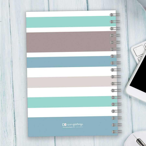 SparkGreetings® Array of Stripes Writing Journal, pastel-colored striped notebook with a spiral binding on a light wooden desk.