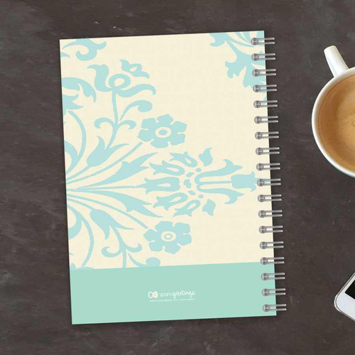 SparkGreetings® Tapestry Pattern Writing Journal, spiral-bound notebook with a light blue floral design on a cream background, placed on a dark wooden surface with a cup of coffee and a smartphone nearby.