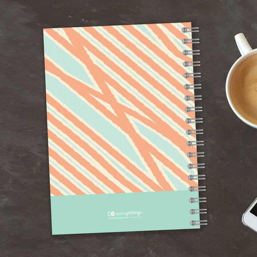 SparkGreetings® Tapestry Pattern Writing Journal, spiral-bound notebook with a vibrant, colorful tapestry-inspired cover design on a dark wooden surface next to a cup of coffee and a smartphone.