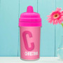 Spark & Spark Double Initial Pink Sippy Cup, a pink children's sippy cup with a large letter C and the name Christina written on it, placed on a white surface with a light blue background and a pink flower in a glass vase.