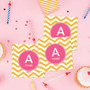 SparkGreetings® Chevron Mustard & Pink Gift Label Set, stationery with yellow and white chevron pattern labels featuring a pink circle and white letter 'A' and the name 'Amelie' on a pink background, alongside party candles and cupcakes.