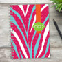 SparkGreetings® Zebralicious Writing Journal, vibrant zebra-stripe patterned cover with pink, blue, white, and orange colors, green 'acb' logo, spiral binding, on a wooden surface with a black pen and green plant nearby.