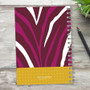 SparkGreetings® Zebralicious Writing Journal, vibrant zebra print design with purple, white, and brown stripes on the cover, yellow polka dot band at the bottom with SparkGreetings logo, spiral-bound notebook on a wooden surface with a black pen nearby and a green plant in the background.