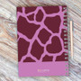 SparkGreetings® Cute Rocks Writing Journal, featuring a purple and pink giraffe-print cover with a spiral binding and a matching pen, placed on a wooden surface.