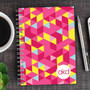 SparkGreetings® Dynamic Ways Writing Journal, colorful geometric pattern cover with pink, yellow, light blue, and red triangles, spiral-bound notebook on wooden desk with a cup of coffee, pen, and smartphone nearby.