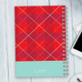SparkGreetings® Criss Cross Writing Journal, red plaid patterned spiral-bound notebook with light blue section at the bottom, placed on a white wooden surface with a partially visible smartphone and computer keyboard.