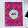SparkGreetings® Criss Cross Writing Journal, pink cover with yellow, white, and purple crisscross patterns, personalized with a blue round monogram and name, placed on a light wooden surface beside a smartphone and keyboard.