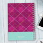 SparkGreetings® Criss Cross Writing Journal, pink and purple plaid patterned cover with a turquoise bottom section and wire binding, placed on a light wooden surface.