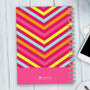SparkGreetings® Style in Stripes Writing Journal, colorful spiral-bound notebook with a vibrant chevron pattern in pink, yellow, orange, and blue on a wooden surface.