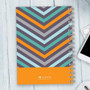 SparkGreetings® Style in Stripes Writing Journal, colorful striped pattern with orange accent at the bottom, spiral-bound notebook on a light wooden desk with a smartphone and keyboard.