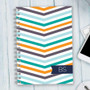 SparkGreetings® Beautiful Colors Writing Journal, a spiral-bound notebook with a vibrant chevron pattern in orange, navy, gray, and teal on the cover, placed on a light wooden surface with a partial view of a smartphone and computer keyboard.