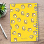 SparkGreetings® Small Swirls Writing Journal, a yellow spiral-bound notebook with a floral swirl pattern on the cover, placed on a wooden desk next to a silver pen and a plant.