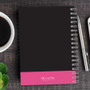 SparkGreetings® Victorian Bold Style Writing Journal, black cover with pink band at the bottom, spiral-bound notebook on wooden desk with pen, coffee cup, and smartphone.