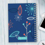 SparkGreetings® Deconstructed Nature Writing Journal, vibrant blue cover with modern abstract floral patterns in red, white, teal, and dark blue, spiral binding, placed on light wooden surface with a white smartphone and keyboard nearby.