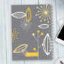 SparkGreetings® Deconstructed Nature Writing Journal, gray cover with yellow and white floral abstract design, spiral-bound notebook on a light wooden desk with a white smartphone and keyboard nearby.