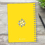 SparkGreetings® Stamped Blossoms Writing Journal, bright yellow notebook with floral stamped design on the cover, spiral binding, and a black pen beside it on a rustic wooden surface.