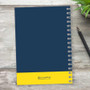 SparkGreetings® Elegant Ribbon Writing Journal, blue hardcover journal with a yellow bottom band and a silver spiral binding, placed on a wooden surface with a black pen and green plant nearby.
