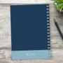 SparkGreetings® Elegant Ribbon Writing Journal, blue hardcover notebook with a spiral binding and a light blue bottom section, placed on a rustic wooden surface with a pen and a green plant nearby.