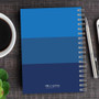 SparkGreetings® Shades of Color Writing Journal, blue gradient cover with spiral binding on wooden desk with pen, coffee cup, and green fruit.