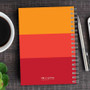 SparkGreetings® Shades of Color Writing Journal, vibrant orange and red spiral notebook placed on wooden desk with a cup of coffee, a pen, and a smartphone.