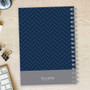 SparkGreetings® Herringbone Mood Writing Journal, navy blue herringbone pattern cover with gray section at the bottom, spiral binding on the right side, placed on a light wooden desk with a keyboard, paper clips, and a clock nearby.