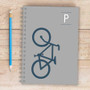 SparkGreetings® Bike Ride Writing Journal, gray spiral-bound journal with a simple bicycle illustration on the cover, placed on a wooden surface alongside a blue pencil.