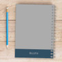 SparkGreetings® Bike Ride Writing Journal, a spiral-bound notebook with a gray cover and blue bottom section, placed on a wooden surface alongside a blue pencil.