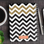 SparkGreetings® Black Chevron Writing Journal, notebook with a black and gold chevron pattern cover on a wooden desk, with a pen, coffee cup, and a small plant nearby.