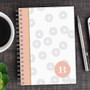 SparkGreetings® Spinning Circles Writing Journal, stylish spiral-bound journal with a white cover featuring gray circular patterns, pink accent stripe, and a monogrammed letter B in a peach circle, placed on a wooden desk with a pen, coffee cup, and phone nearby.