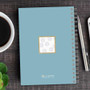 SparkGreetings® Spinning Circles Writing Journal, light blue spiral notebook with a decorative white square center featuring gray circles, on a wooden desk with a silver pen, coffee cup, and green succulent plant.