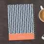 SparkGreetings® Triangles & Glitter Writing Journal, a notebook with a blue and white triangle pattern cover and a coral bottom band, placed on a dark wooden surface next to a cup of coffee and a smartphone.