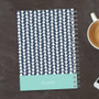 SparkGreetings® Triangles & Glitter Writing Journal, a navy blue and white geometric patterned notebook with a spiral binding, placed on a dark wooden surface next to a cup of coffee and a smartphone.
