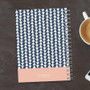 SparkGreetings® Triangles & Glitter Writing Journal, geometric patterned journal with navy and white triangles and a pink band at the bottom, placed on a dark wooden surface with a white coffee cup and smartphone nearby.