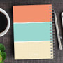 SparkGreetings® Three Stripes Writing Journal, colorful notebook with pastel-colored horizontal stripes on the cover, spiral binding, placed on a dark wooden desk with a coffee cup and pens.