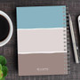 SparkGreetings® Three Stripes Writing Journal, a spiral-bound notebook with a color-blocked cover in blue, white, and brown, placed on a wooden desk alongside a cup of coffee, a silver pen, and a mobile phone.