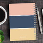 SparkGreetings® Three Stripes Writing Journal, colorful notebook with horizontal color blocks in pink, blue, and beige, spiral binding, on a wooden desk with a pen, coffee, and plant nearby.