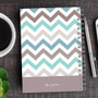 SparkGreetings® Colorful Chevrons Writing Journal, with a cover design featuring pastel-colored chevron patterns and a gray bottom section, placed on a wooden desk alongside a cup of coffee, a silver pen, and a smartphone.