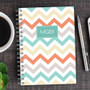 SparkGreetings® Colorful Chevrons Writing Journal, patterned spiral-bound notebook with pastel chevron design in orange, teal, beige, gray, and white on a wooden desk with a cup of coffee, a silver pen, and a smartphone.
