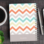 SparkGreetings® Colorful Chevrons Writing Journal, with a vibrant cover design featuring multicolored chevron patterns in shades of orange, teal, yellow, and gray on a wooden desk.