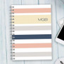 SparkGreetings® Array of Stripes Writing Journal, a spiral-bound notebook with horizontal pastel-colored stripes and the initials 'MGB' on the cover placed on a light wooden surface.
